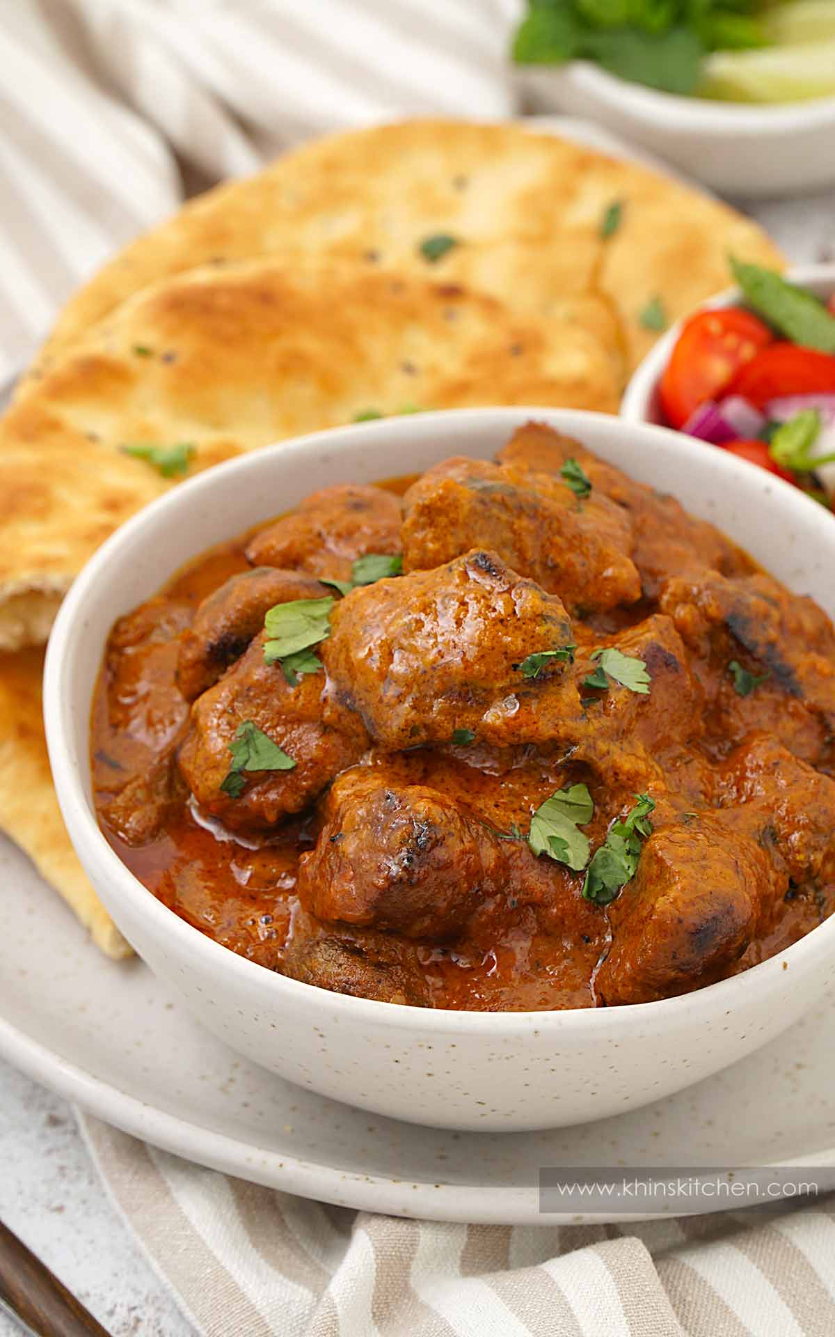 A white bowl containing creamy lamb curry with naan and onion salad on the side. 