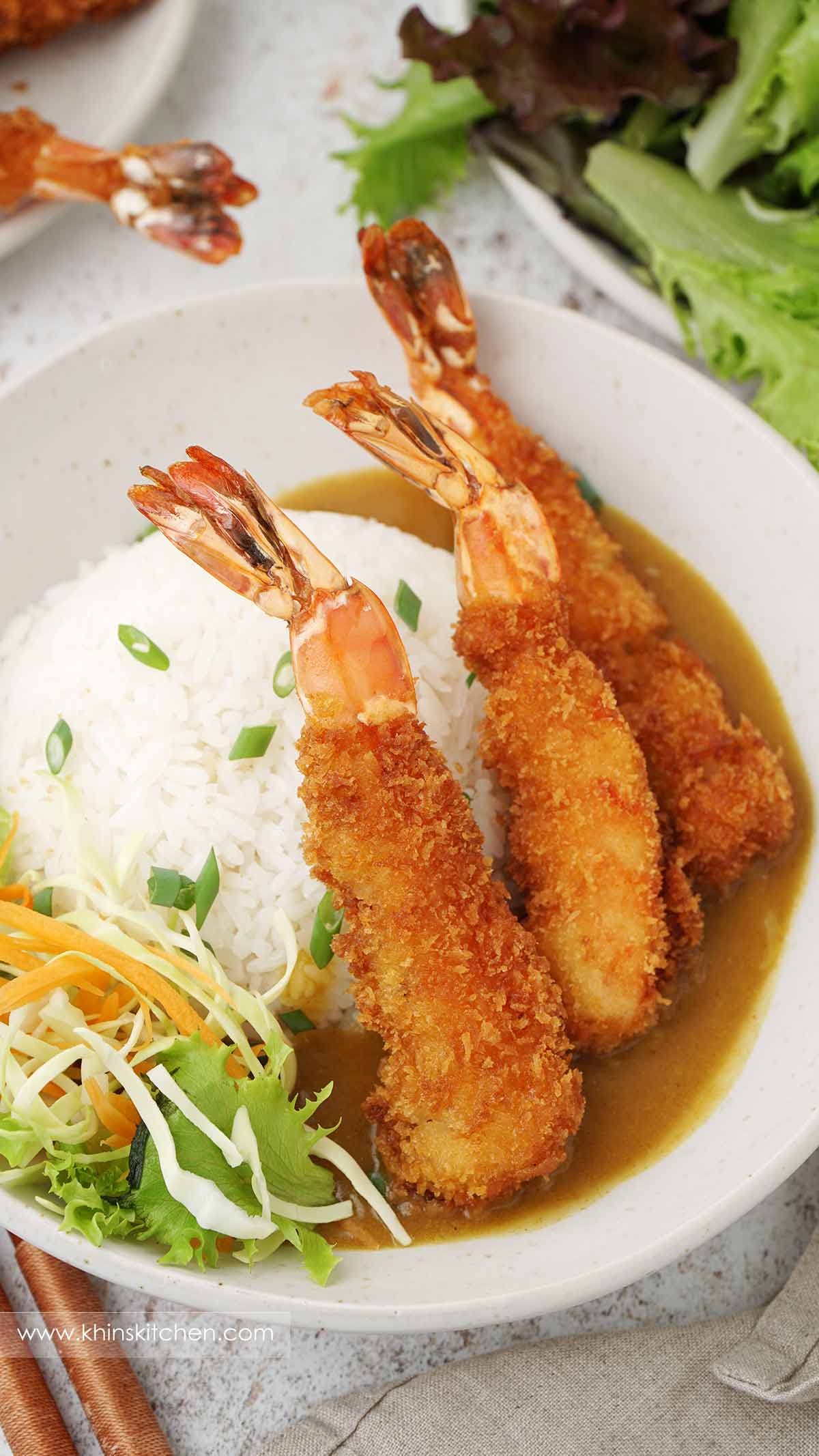 A grey bowl containing fried katsu prawns with rice and curry sauce. Shredded cabbage and carrot on the side.