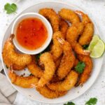 A white plate containing fried coconut prawns with dipping sauce and lime wedges on the side.