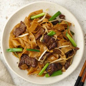 A white plate containing, flat rice noodles with beef, spring onions and bean sprouts.