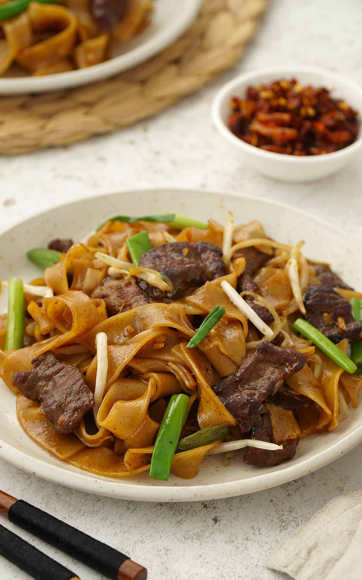 A white plate containing stir fried wide rice noodles with beef, bean sprouts, and spring onions. 