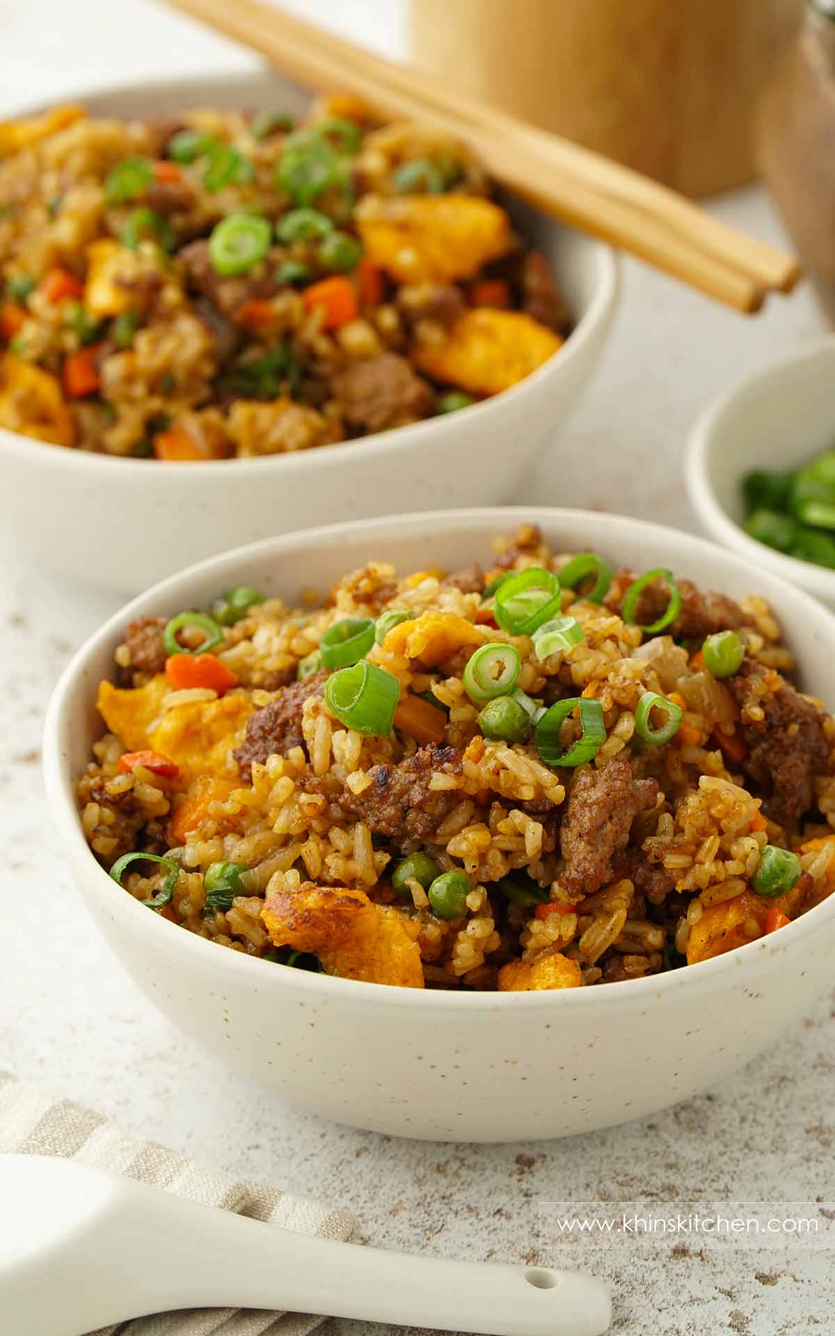 A white bowl containing Chinese fried rice with minced beef, spring onions, scrambled egg, carrots, and green peas. 