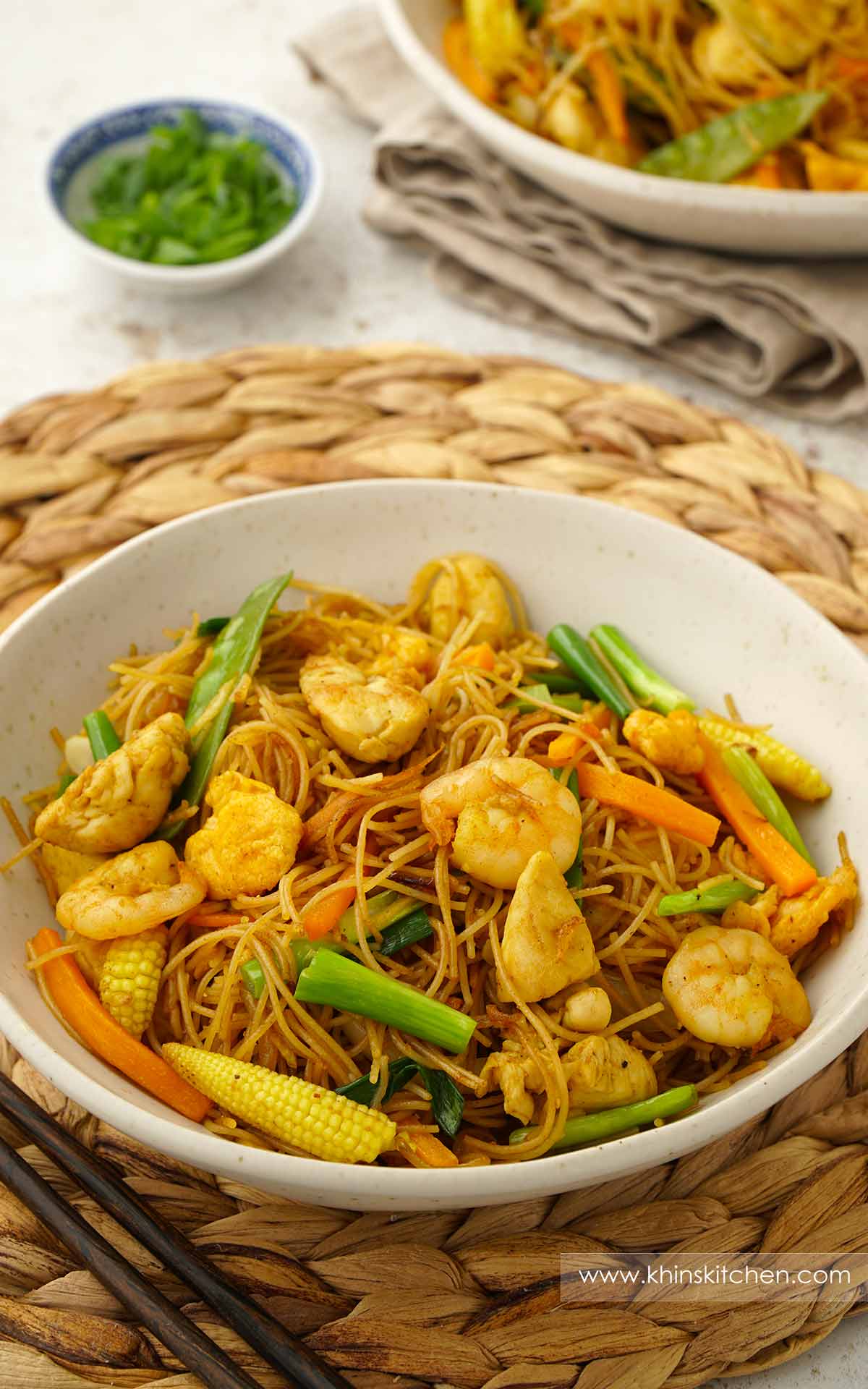 A white bowl containing fried rice noodles with prawns, chicken, spring onions, carrot, and baby corn.