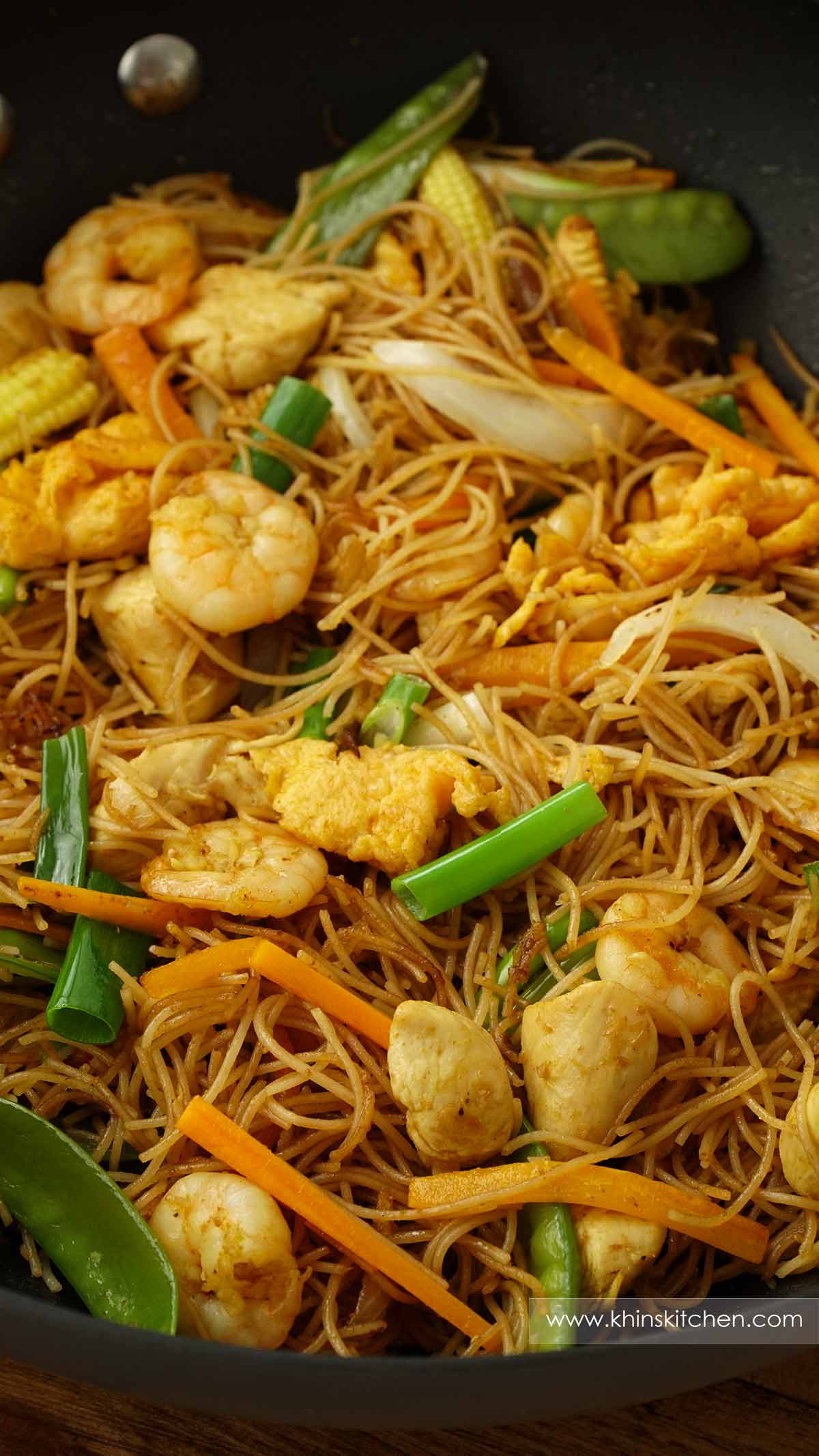 A black wok containing, stir fried Singapore-style mei fun noodles with eggs, prawns, chicken and vegetables.