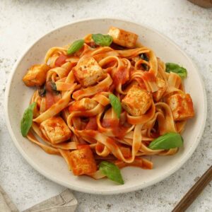 A white plate containing, pasta in tomato sauce with tofu, fresh basil and grated cheese.