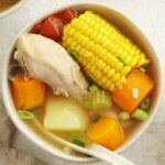 A white bowl containing, chicken soup with corn, carrot, potato, and toamtoes.
