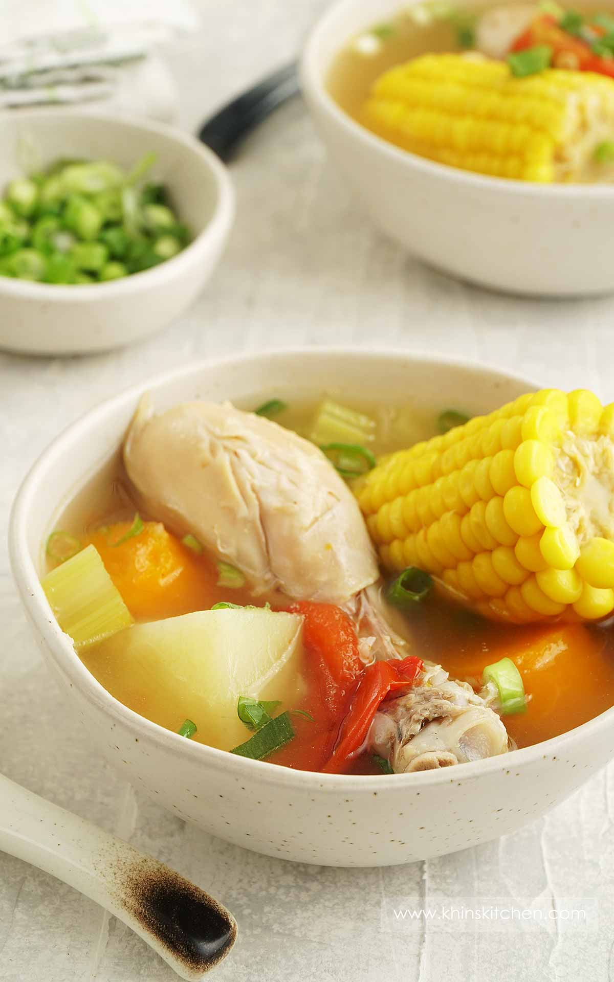 A ceramic bowl containing, soup with chicken drumstick, sweetcorn, potato, tomato, carrot, and celery. 