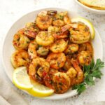 A grey plate containing pan-fried prawns with garlic butter sauce, garnished with parsley and lemon wedges.