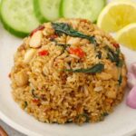 A white plate containing Thai fried rice with chicken, red chillies, and basil leaves. Cucumber, onion, and lemon wedges on the side.
