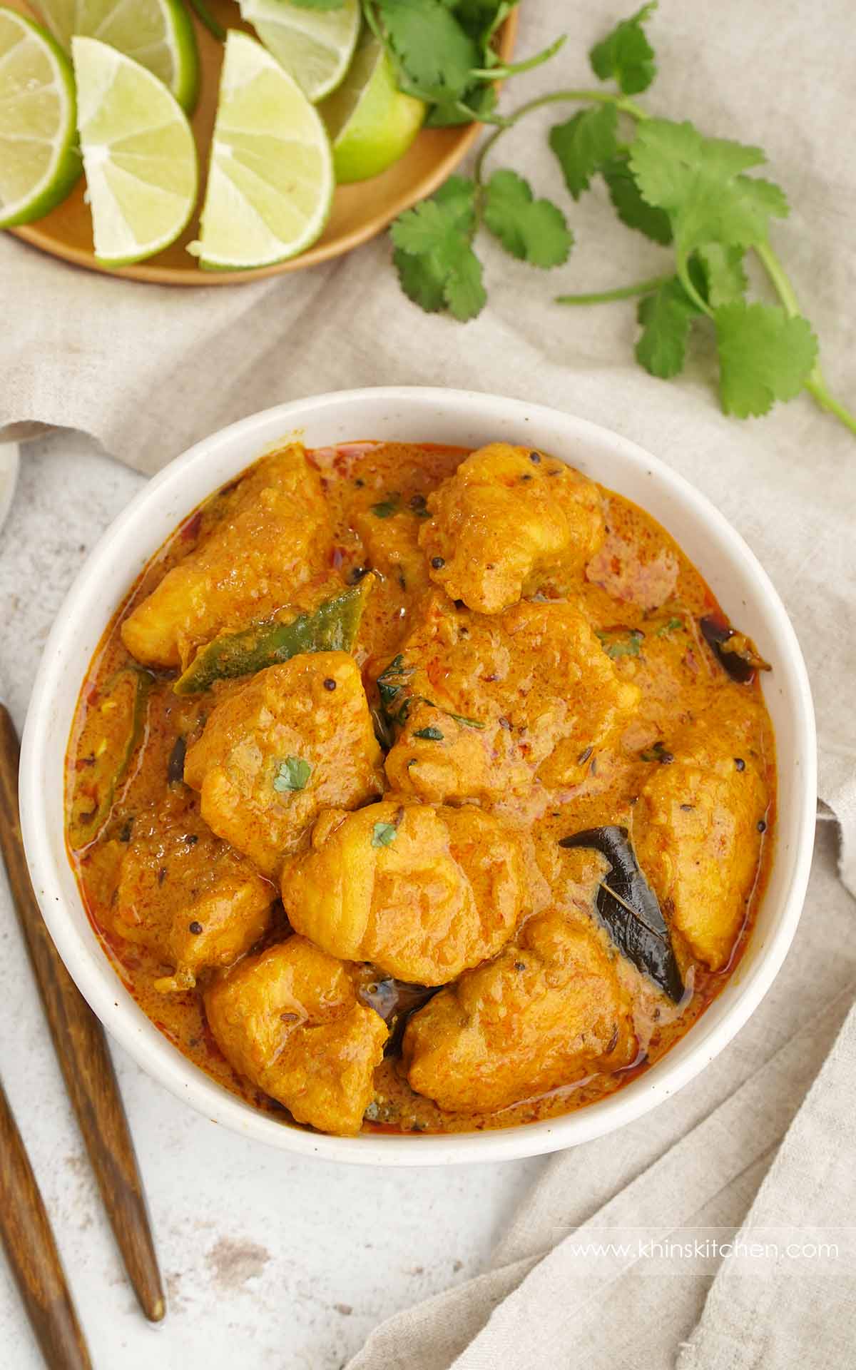 A white bowl containing, fish pieces cooked in, creamy curry sauce with curry leaves and green chillies. 