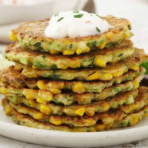 A white plate containing, layers of courgette and sweetcorn fritters, topped with yoghurt dipping sauce.