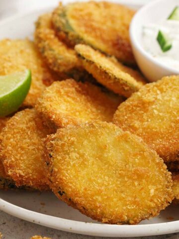 A white plate containing, panko coated fried courgette slices with yoghurt dipping sauce on the side.