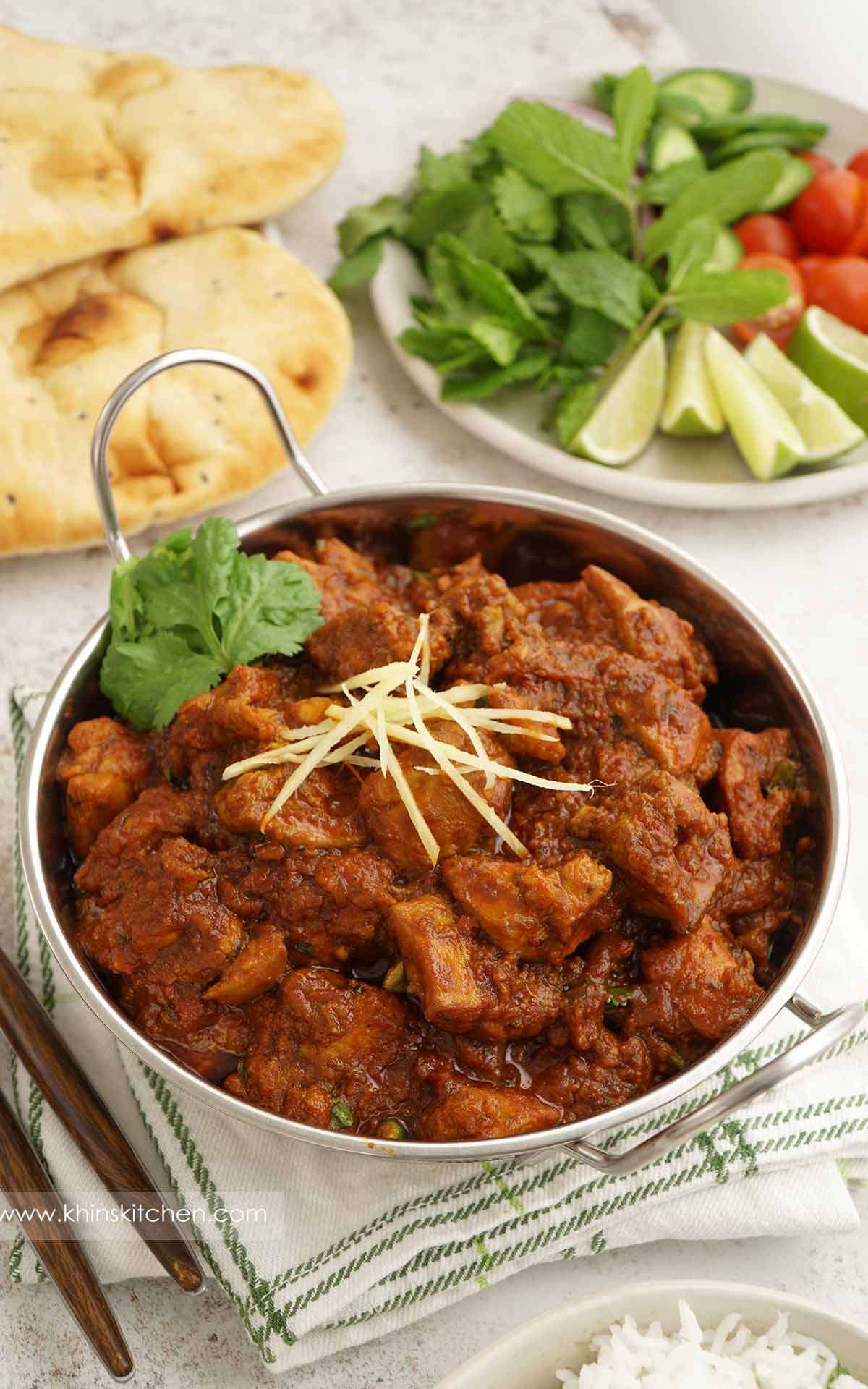 A steel bowl containing spicy chicken curry topped with ginger slices and coriander.