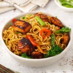 A white bowl containing teriyaki salmon cubes with vegetables, noodles, and sesame seeds.