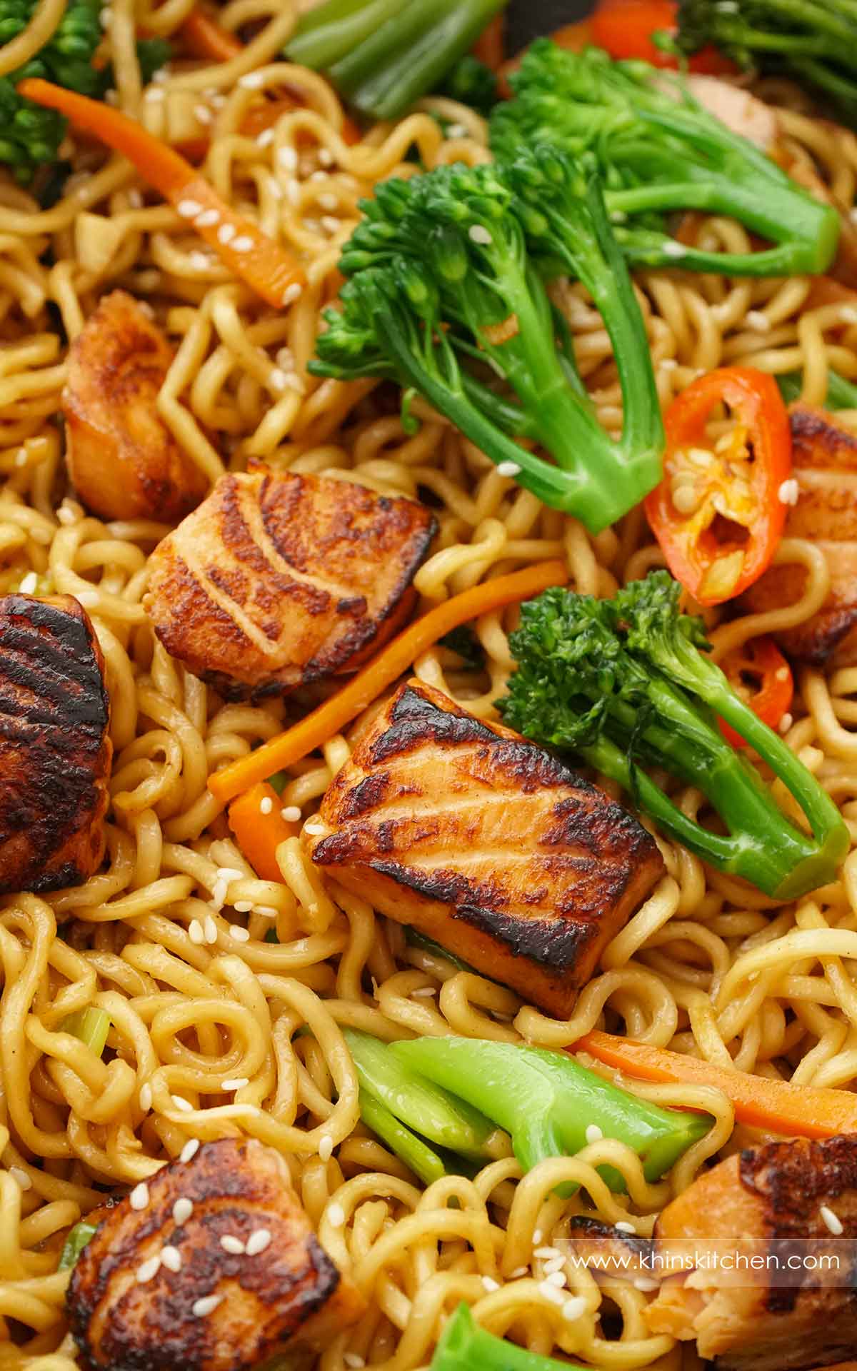 A close up photo of ramen noodles with salmon, broccoli, carrot, chilii pepper, and sesame seeds. 