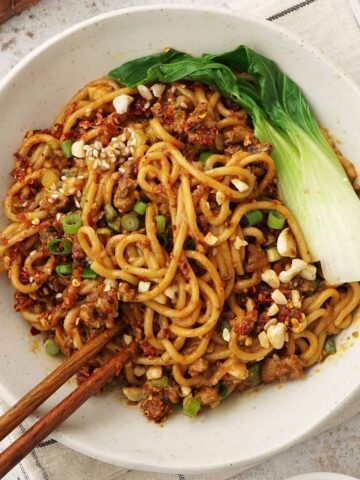 A white bowl containing, spicy Sichuan dan dan noodles with minced meat, crushed peanuts, sesame seeds, and blanched vegetables.