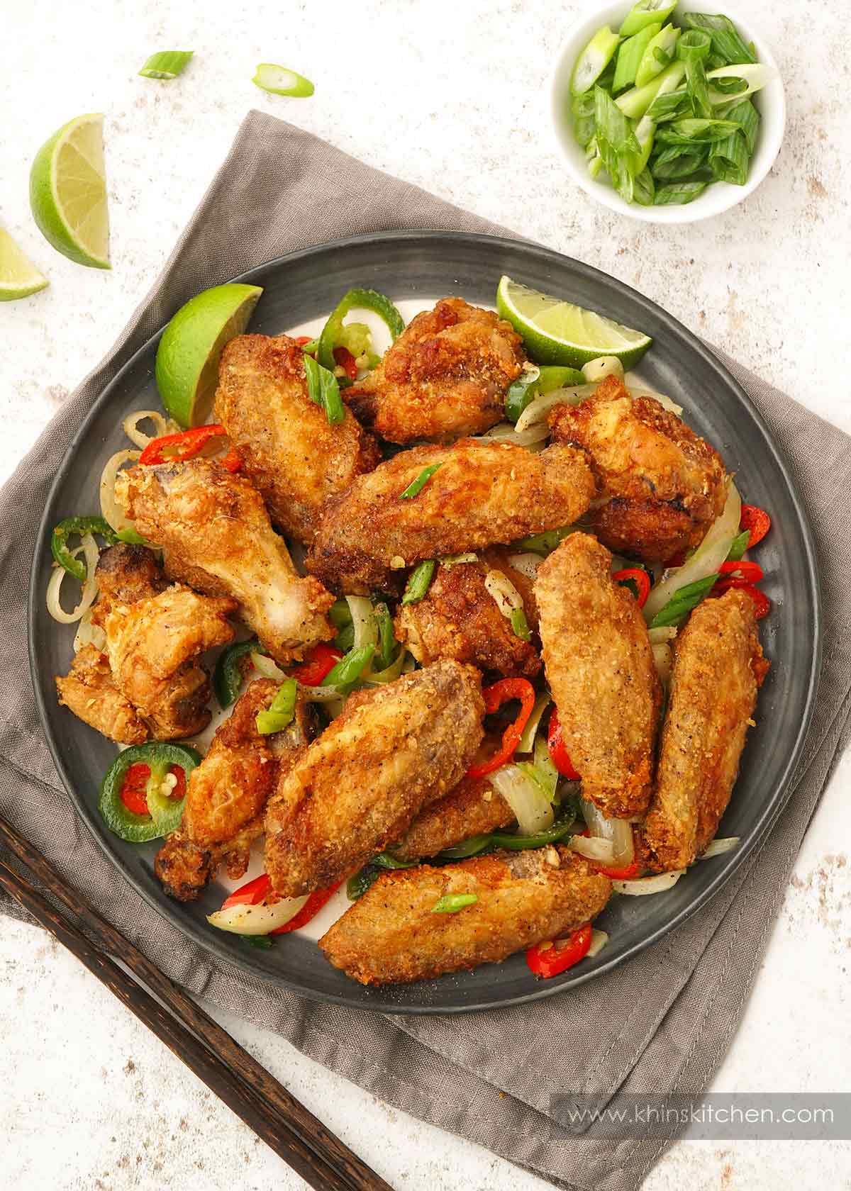 A grey plate containing crispy fried golden colour chicken wings with vegetables, salt, and pepper seasonings. 