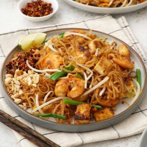 A white plate containing, stir fried Thai thin rice noodles with prawns, bean sprouts, spring onions, crushed peanuts, and chilli flakes.