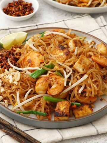 A white plate containing, stir fried Thai thin rice noodles with prawns, bean sprouts, spring onions, crushed peanuts, and chilli flakes.
