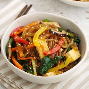 A white bowl containing Japchae noodles with colourful vegetables, tofu, and sesame seeds.