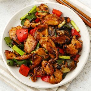 A white plate containing stir fry chicken with bell peppers, spring onions, garlic, and fermented black beans.