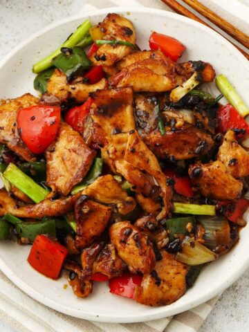 A white plate containing stir fry chicken with bell peppers, spring onions, garlic, and fermented black beans.