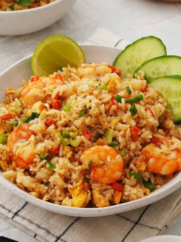 A white bowl containing, Thai fried rice with prawns, eggs, chillies, and spring onions. Garnished with cucumbers and lime wedges.