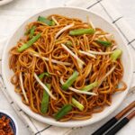 A white plate containing, fried brown chow mein noodles with bean sprouts and green onions.