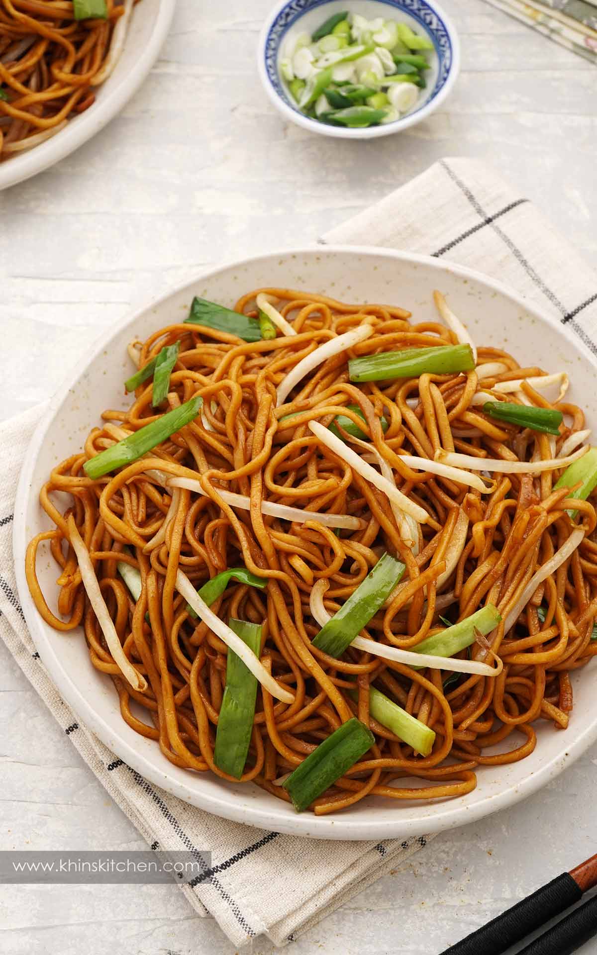 A white plate containing brown colour stir fried noodles with bean sprouts and spring onions. 