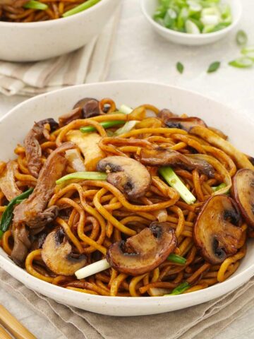 A white bowl containing stir-fried noodles, mushrooms, and spring onions.