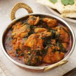 A metal bowl containing, Indian spinach curry with chicken thigh pieces.