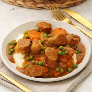 A white plate containing curried sausages with carrots, green peas and mashed potato.