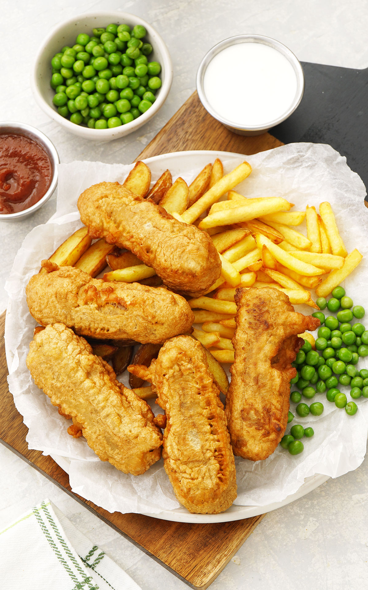 A white plate containing fried batter coated sausages, fries, and green peas.