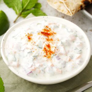 A white bowl containing Indian yoghurt sauce with vegetables and garnished with mint leaves and ground spices.