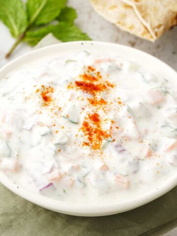 A white bowl containing Indian yoghurt sauce with vegetables and garnished with mint leaves and ground spices.