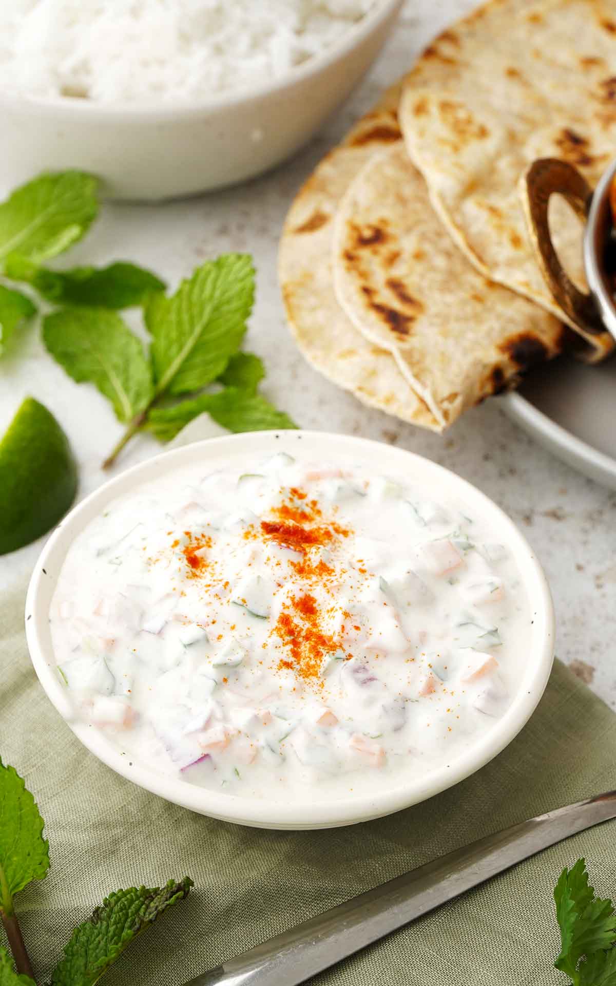 A white bowl containing Indian yoghurt sauce with vegetables and garnished with ground spices. Naan bread and mint leaves on the side.