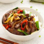 A white bowl containing stir fry beef slices with brown glossy sauce and vegetables.