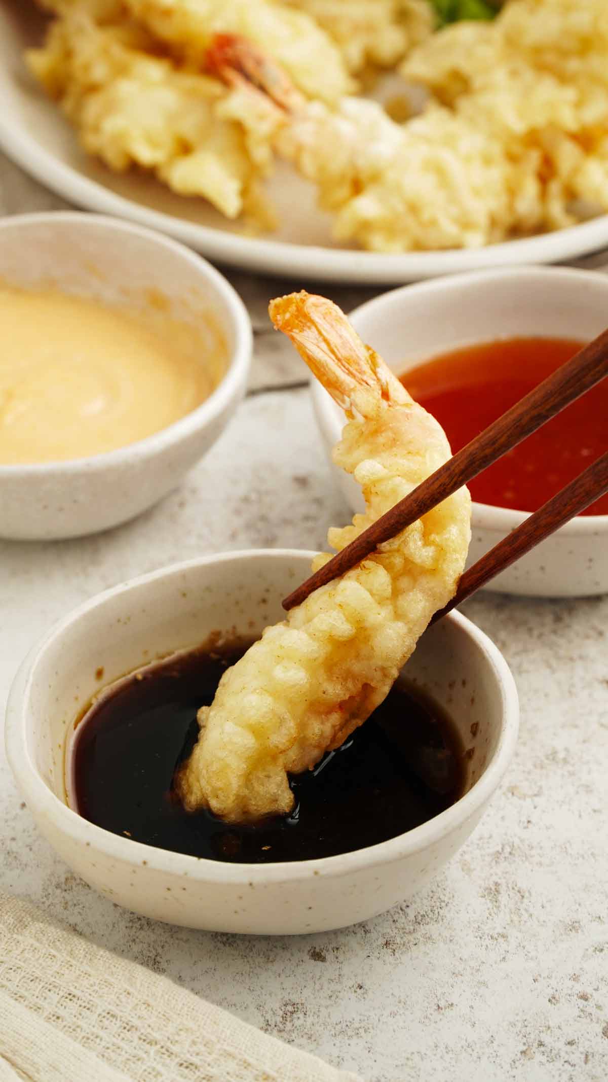 A pair of chopstick holding a fried prawns, dipped in a soy dipping sauce.