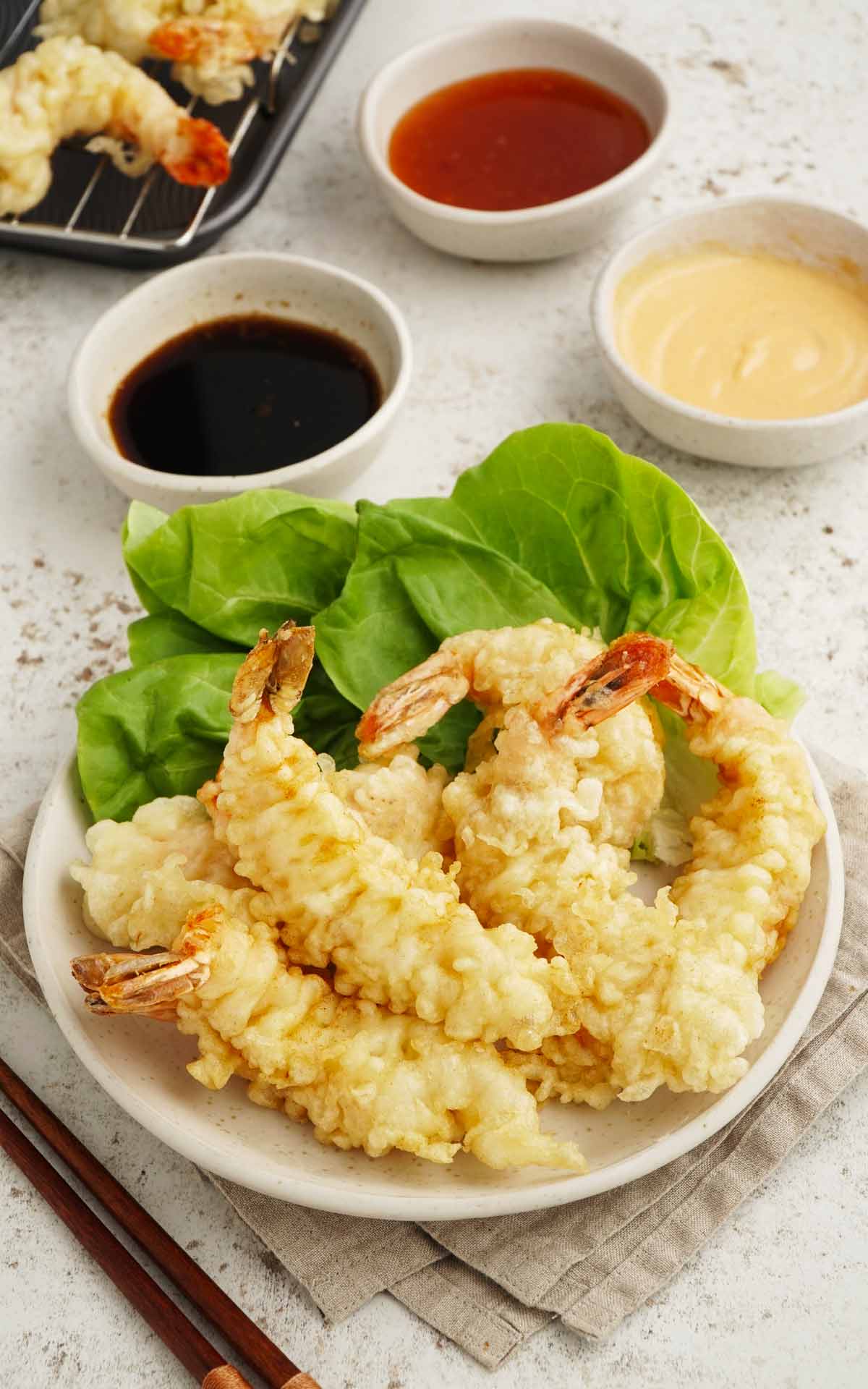 A white plate containing fried crispy Japanese tempura battered prawns, garnished with salad leaves, and dipping sauces on the side. 