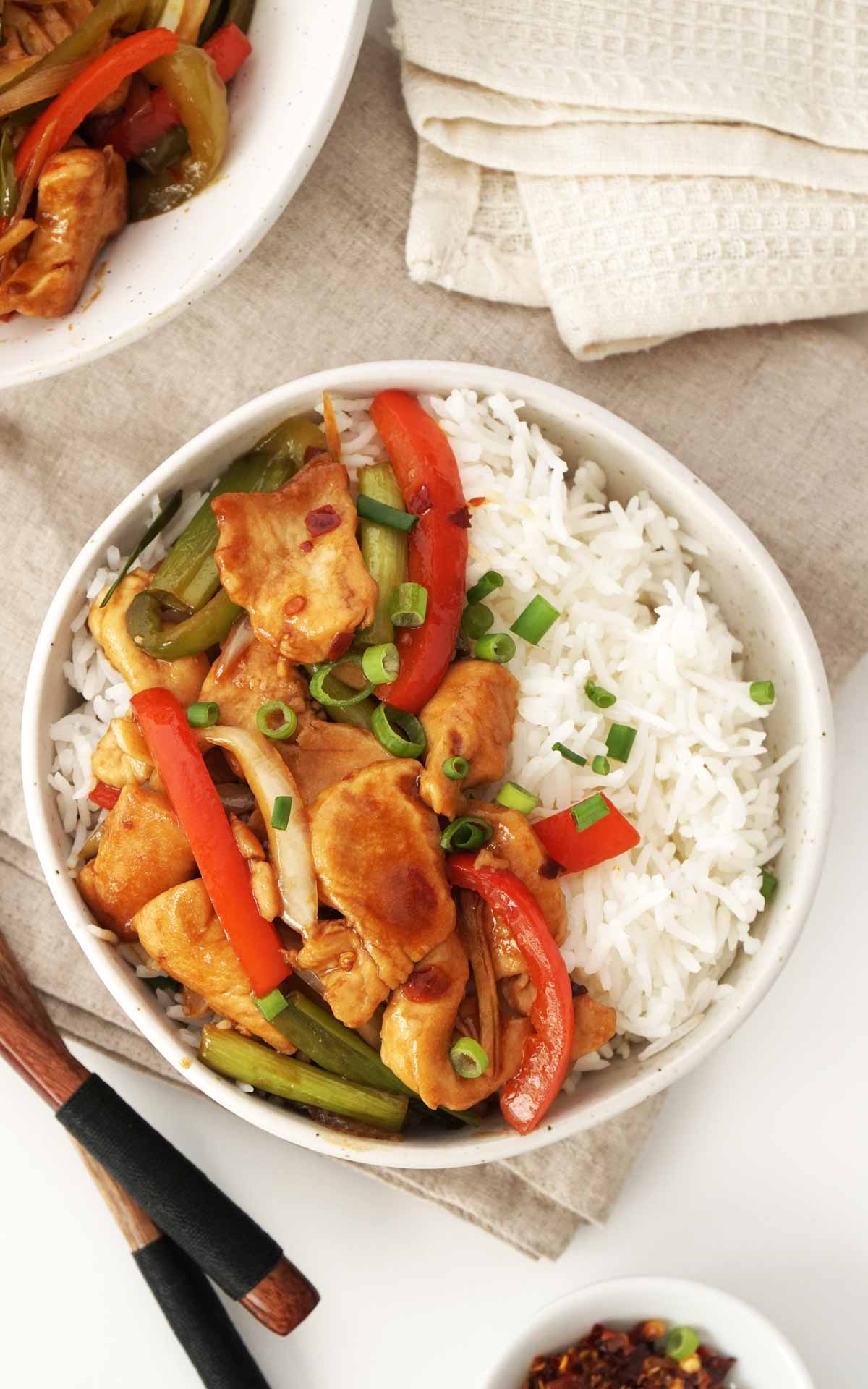 A rice bowl containing stir fried chicken with bell peppers, spring onions, and hoisin sauce. 