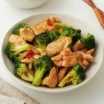 A white bowl containing, stir-fried chicken pieces with vibrant green broccoli, and red chilli slices.