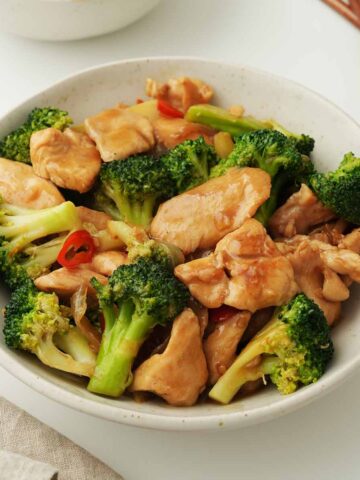 A white bowl containing, stir-fried chicken pieces with vibrant green broccoli, and red chilli slices.