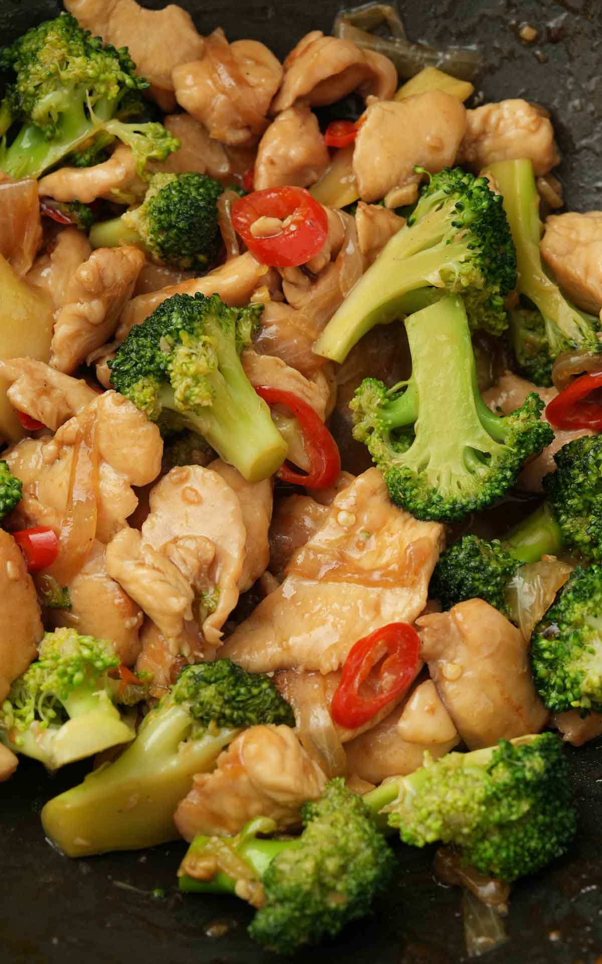 A black wok containing chicken slices, broccoli, red chilli slices, with glossy sauce. 