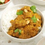 A white bowl containing creamy lamb curry with rice and garnished with almond flakes and coriander.