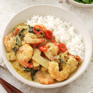 A white ceramic bowl containing, white rice, prawns, bamboo shoots, Thai basil, red chilli slices, and creamy curry sauce.