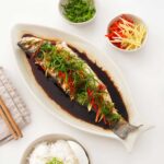A white plate containing Chinese-style whole steamed fish with ginger, chilli, and scallions.
