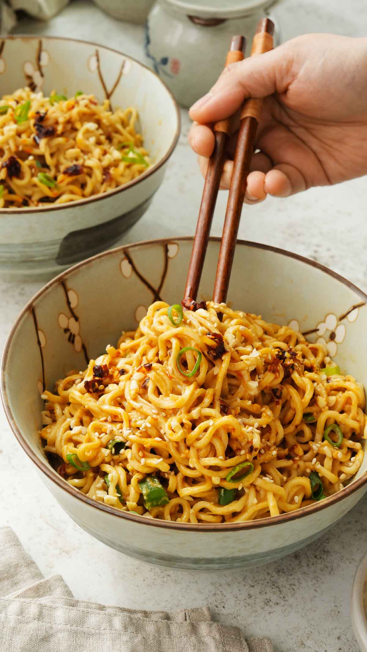 Noodle bowl containing ramen noodles with chilli peanut sauce. A pair of chopsticks holding the noodles. 