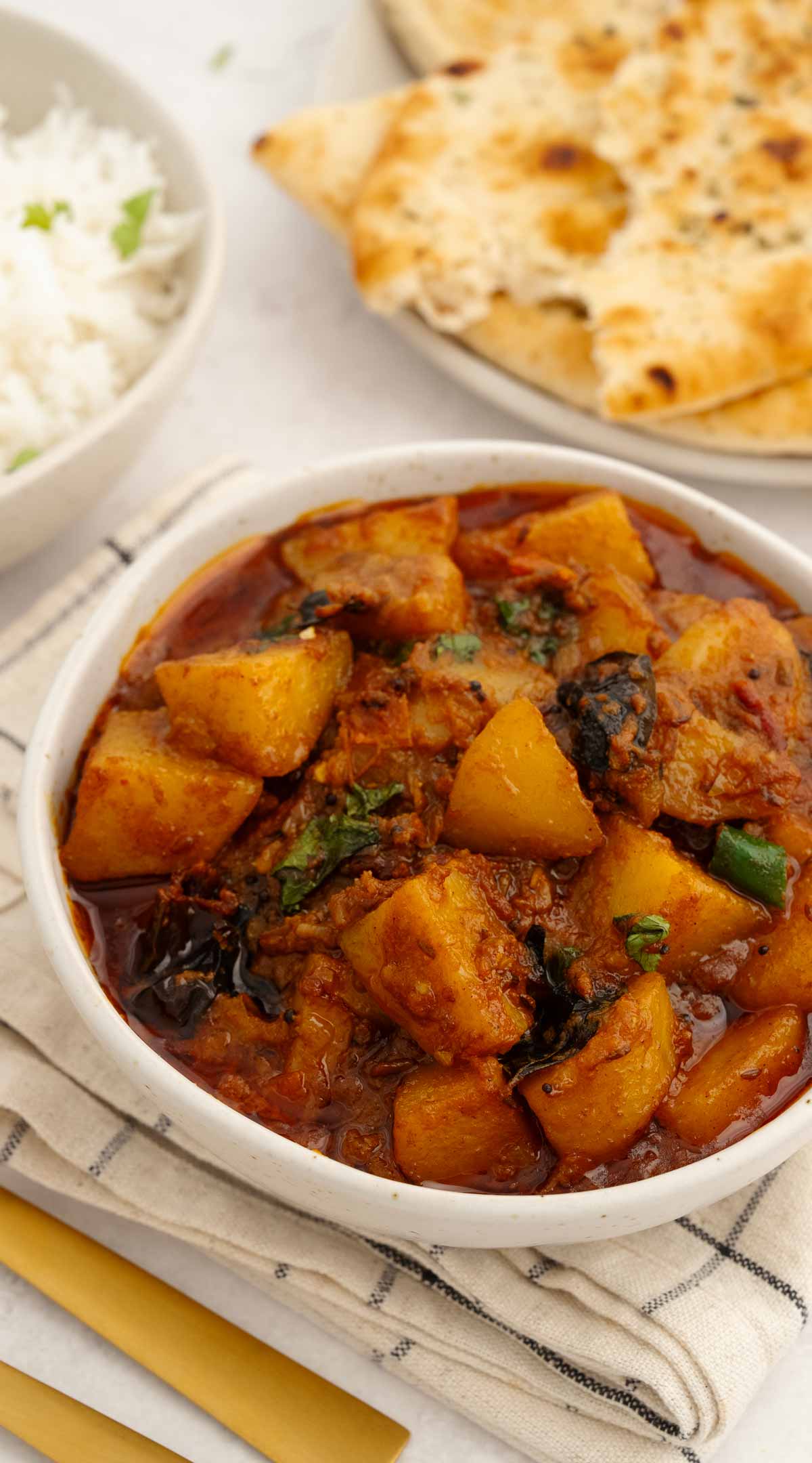 A white bowl containing diced potatoes in onions and tomatoes curry sauce. Garlic naan and rice on the side. 