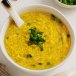A white bowl containing, creamy yellow colour egg drop soup with a white ceramic spoon inside.