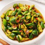 A white plate containing, cucumber pieces with seasoning sauces and red chillies.